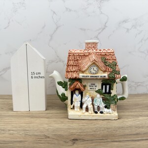 Könnte beinhalten: Eine dekorative Teekanne in Form eines Häuschens mit Strohdach, Uhr und dem Schriftzug "LILLEY CRICKET CLUB". Die Teekanne ist mit Figuren und einem Hund verziert. Das weiße hausförmige Objekt ist 15 cm hoch.
