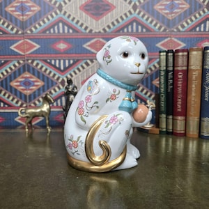 May include: A white ceramic monkey figurine featuring delicate floral patterns in pink, green, and orange. The monkey wears a light blue collar with tassels, has a metallic gold tail, and holds a small peach-colored sphere in its hands.