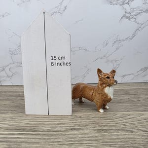 Puede incluir: Una figura de corgi marrón y blanco está junto a un objeto de madera blanco en forma de casa. La casa mide 15 cm de alto y tiene las medidas de 6 pulgadas escritas. El corgi tiene un diseño detallado y está sobre una superficie de madera.