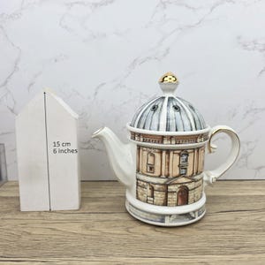 Op de afbeelding: Een witte keramische theepot met een gebouwontwerp, een goudkleurig deksel en een gebogen handvat. De theepot is ongeveer 15 cm hoog. Het gebouwontwerp heeft beige en bruine tinten, met een koepelvormig deksel en een tuit.