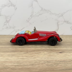 Peut inclure: Une voiture jouet rouge vintage avec des roues noires et un pare-brise argenté. La voiture classique a un pare-brise bleu et un design détaillé, mettant en valeur une esthétique rétro. La voiture jouet est en métal et repose sur une surface en bois.