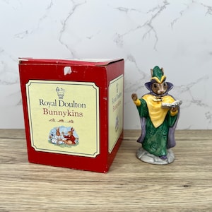 以下が含まれることがあります： ロイヤル・ダルトン・バニキンズのフィギュアで、緑と紫のローブを着て、黄色のサッシュをつけたウサギが本を持っています。フィギュアは緑と紫のデザインの台座の上に立っています。フィギュアは、赤い箱に入っていて、箱には「ロイヤル・ダルトン・バニキンズ」と書かれた白いラベルが付いています。