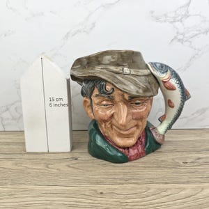 Op de afbeelding: Een keramische mok in de vorm van een mannenhoofd, met een bruine hoed en groene jas, met een vissenhandvat. De mok is ongeveer 15 cm hoog. Het vissenhandvat heeft rode en witte accenten.