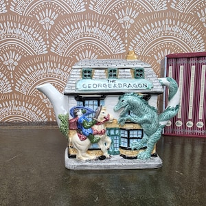 May include: A ceramic teapot shaped like a building with a sign reading THE GEORGE & DRAGON. The front features a figure on a white horse facing a green dragon. The roof is grey with a small chimney, and the handle is shaped like a green dragon tail.