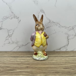 Op de afbeelding: Een keramieken konijnfiguur met een gele vest en een paarse jas, die een zakhorloge vasthoudt. Het konijn staat op een grasveldje. Het beeldje is gedetailleerd beschilderd en heeft een klassiek ontwerp.