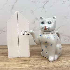 May include: A white ceramic cat-shaped teapot with blue floral accents and green eyes. The teapot is approximately 15 cm tall. A white wooden house shape stands next to the teapot.