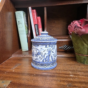 Op de afbeelding: Een blauw-witte keramische pot met deksel, met een handgeschilderd ontwerp van bloemen en architecturale elementen. De pot is cilindrisch en staat op een houten oppervlak. De pot is ongeveer 13 cm hoog.