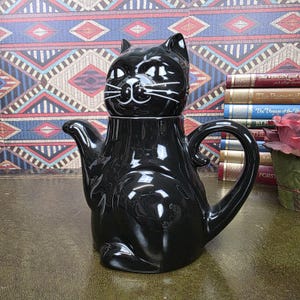 Op de afbeelding: Een glanzende zwarte keramische theepot in de vorm van een zittende kat. De theepot heeft een deksel in de vorm van een kattenkop met wit geschilderde ogen, neus en snorharen. Het heeft een gebogen handvat aan de rechterkant en een tuit aan de linkerkant.