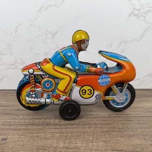 Pode incluir: Uma motocicleta de brinquedo vintage de lata com um motociclista em um traje azul e amarelo. A motocicleta é laranja e azul com uma roda branca e preta. A motocicleta tem o número "93" no lado e as palavras "Orient Exports" na parte de trás.