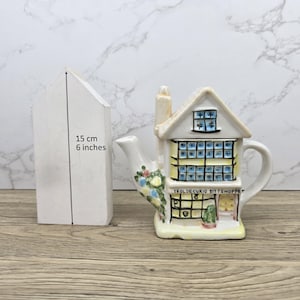 Puede incluir: Una tetera de cerámica con forma de casa de dos pisos con chimenea, ventanas y puerta. La tetera está pintada en crema, amarillo y azul. El texto "YEOLDECURIO SITTSHOPPE" es visible. Un objeto blanco en forma de casa mide 15 cm.