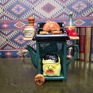Peut inclure: Une théière en céramique en forme de barbecue au charbon, avec un corps vert, un couvercle noir et un sac jaune de charbon Little Devil. Le dessus porte un poulet rôti et des accompagnements, avec une petite roue en bois sur la base avant.