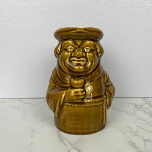 May include: A brown ceramic pitcher shaped like a monk holding a cup. The monk has a smiling face and a brown robe.