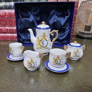 Könnte beinhalten: Ein Teeservice aus weißem Porzellan mit Goldrand und blauen Akzenten, das an das Goldene Thronjubiläum von Königin Elizabeth 1952-2002 erinnert. Das Set umfasst eine Teekanne, eine Zuckerdose mit Deckel, ein Milchkännchen und zwei Tassen.