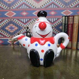 Może przedstawiać: Ceramiczny czajniczek w kształcie klauna z białą podstawą, czerwonymi kropkami i niebieskimi kropkami. Klaun ma czerwony nos, namalowany uśmiech, czarne buty i czarny kapelusz. Czajniczek posiada zakrzywiony uchwyt oraz dzióbek wystający z boku.