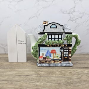 May include: A ceramic teapot shaped like an antique shop building with a white spout and handle. The facade features the text "ANTIQUE'S", "BRIC-A-BRAC", and "BOOKS FOR SALE". A white measuring tool indicates a height of 6 inches next to the teapot.
