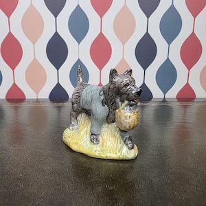 May include: A ceramic figurine of a black dog wearing a blue jacket and carrying a woven basket. The dog stands on a patch of yellow grass. The figurine has a glossy finish and is set against a patterned background.