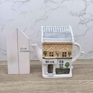 May include: A white ceramic teapot shaped like a bank building, with a gray roof and brick facade. The teapot has a handle and spout, and the word "BANK" is displayed on the front. A white house-shaped object, 6 inches tall, is next to the teapot.