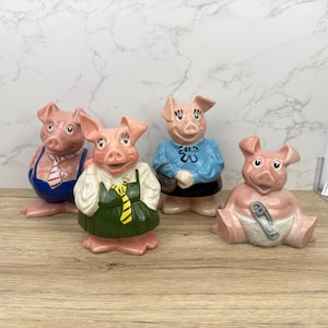 May include: Four ceramic piggy banks, each with a different outfit. One is wearing a blue and white striped shirt with a red tie, another is wearing a green dress with a yellow tie, another is wearing a blue dress with a pink bow, and the last one is wearing a white diaper with a safety pin.