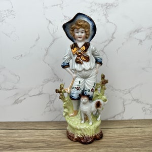 May include: A porcelain figurine of a boy wearing a blue and white sailor outfit and a blue hat, holding a stick and standing next to a white and brown dog. The figurine is on a green and brown base.