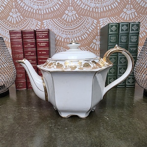 May include: A white porcelain teapot with a hexagonal body and a lid. The teapot features gold trim along the edges, handle, and spout, with a decorative gold floral pattern encircling the upper section. It stands on small, integrated feet.
