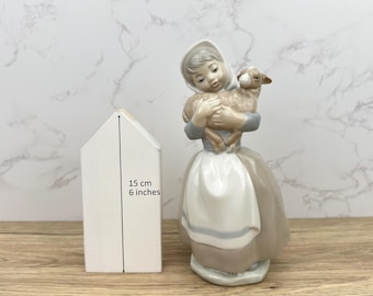 May include: A ceramic figurine of a girl in a bonnet and dress holding a lamb. The figurine is in shades of grey, white, and brown. A white wooden house shape is next to the figurine, measuring 15 cm tall.