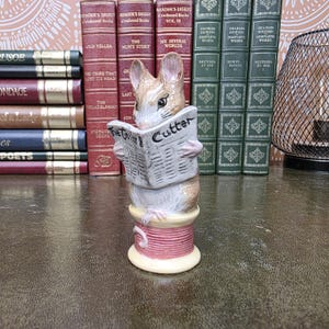 Peut inclure: Une figurine en céramique représentant une souris brune et blanche assise sur une bobine de fil rose. La souris tient un journal ouvert avec le texte The Tailor & Cutter imprimé sur la première page. La figurine est peinte aux couleurs réalistes.