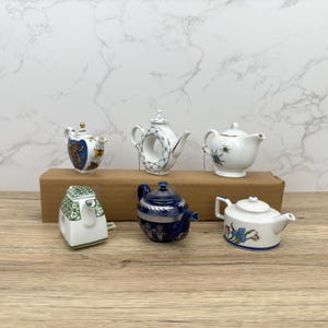 Puede incluir: Seis teteras de porcelana en miniatura en varios colores y dise&ntilde;os. Las teteras est&aacute;n dispuestas sobre una superficie de madera.