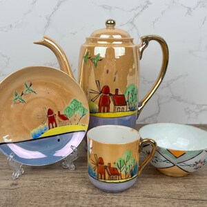 May include: A vintage tea set with a gold trim and a windmill and house design. The set includes a teapot, a cup, a saucer, and a creamer.
