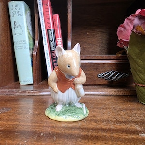 May include: A ceramic mouse figurine wearing a red waistcoat and holding a cane, standing on a patch of green grass. Books and a decorative flower are in the background. The figurine is a collectible item.
