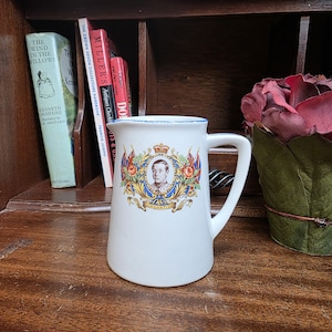 Może przedstawiać: Biały ceramiczny dzbanek z uchwytem, z portretem ukoronowanego mężczyzny i napisem "CORONATION". Dzbanek ma niebieską obwódkę i jest ozdobiony flagami i elementami kwiatowymi. Jest wystawiony na drewnianej powierzchni.