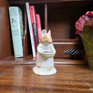 Puede incluir: Figura de cerámica de un ratón con un vestido y sosteniendo un cuenco pequeño. El ratón tiene pelaje marrón claro y viste un vestido blanco con mangas a rayas verdes. La figura mide aproximadamente 8 cm de altura.