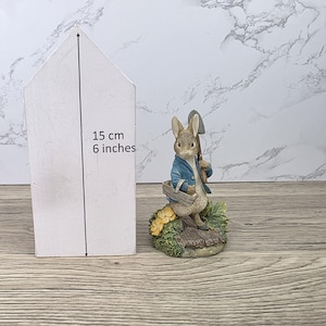 Puede incluir: Figura de un conejo con chaqueta azul, sosteniendo una pala y una cesta, sobre una base con flores y vegetación. Una casa de madera blanca mide 15 cm (6 pulgadas) de alto.