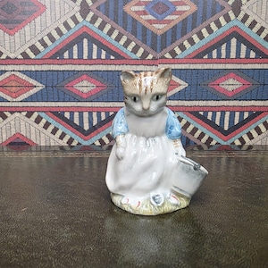 May include: A ceramic figurine of a cat wearing a blue and white dress, holding a small bucket. The cat has brown and white fur and is standing on a patch of grass with small flowers. The background has a colorful geometric pattern.