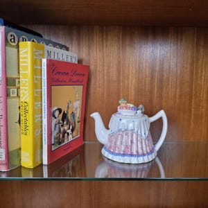 Pode incluir: Uma chaleira de cerâmica branca com um design decorativo rosa e azul. A tampa apresenta um arranjo de frutas e flores. Livros, incluindo um manual do colecionador, são visíveis no fundo.