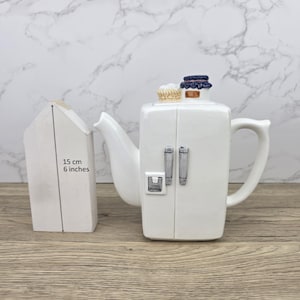 Pode incluir: Uma chaleira de cerâmica branca em forma de geladeira, com alças prateadas e uma pequena cesta e pote em cima. Um objeto branco em forma de casa está ao lado da chaleira, com o texto "15 cm 6 polegadas".