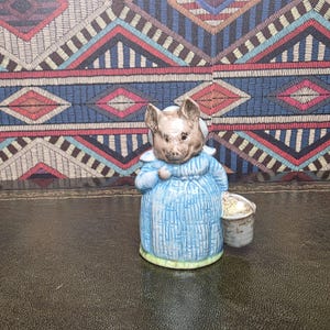 Può includere: Una statuetta in ceramica di un maiale con un vestito blu e un cappello bianco. Il maiale tiene un piccolo secchio. La statuetta ha un viso e orecchie marrone chiaro. L'abito ha linee verticali.