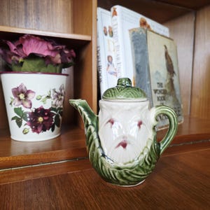 Könnte beinhalten: Eine Vintage-Teekanne aus Keramik mit grünem Griff, Ausguss und Blattdetails. Der Korpus der Teekanne ist weiß mit roten floralen Details. Ein passender grüner Deckel sitzt oben drauf.