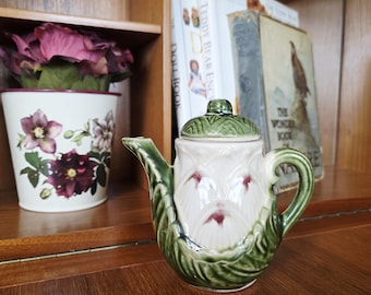 Puede incluir: Una tetera de cerámica vintage con asa, boquilla y detalles de hojas verdes. El cuerpo de la tetera es blanco con detalles florales rojos. Una tapa verde a juego se encuentra encima.