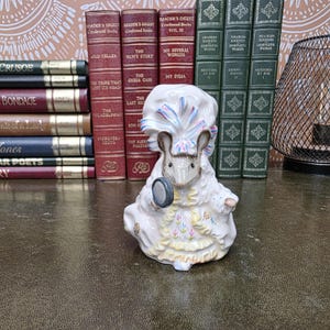 Könnte beinhalten: Eine Porzellanfigur einer Maus in aufwendiger viktorianischer Kleidung, bestehend aus einer gerüschten Haube mit blauen und rosa Bändern, einem hochgeschlossenen Kleid mit gelbem Saum und einer kleinen, runden, dunkel gefärbten Lupe in der Hand.