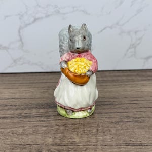 May include: A gray and white porcelain figurine of a squirrel wearing a pink dress and holding a basket of yellow nuts. The figurine is standing on a green base.