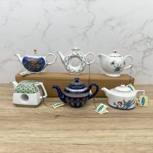 Puede incluir: Seis teteras de porcelana en miniatura con varios dise&ntilde;os. Las teteras son blancas con detalles azules, verdes y dorados. Est&aacute;n dispuestas sobre una superficie de madera.