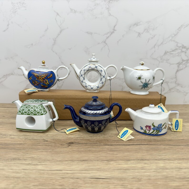 Puede incluir: Seis teteras de porcelana en miniatura con varios dise&ntilde;os. Las teteras son blancas con detalles azules, verdes y dorados. Est&aacute;n dispuestas sobre una superficie de madera.