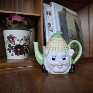 May include: A whimsical ceramic teapot with a corn cob design, featuring a smiling face with blue eyes and rosy cheeks. The teapot has a green handle, spout, and accents. A decorative floral pot and vintage books are in the background.