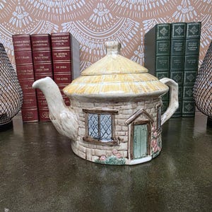 May include: A ceramic teapot shaped like a cottage with a thatched roof, stone-textured walls, a green door, and blue-paned windows. The teapot features a curved spout and a handle, with small pink floral details at the base of the structure.