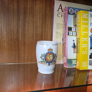 May include: A white ceramic mug featuring a royal crest and portraits of King George VI and Queen Elizabeth. The mug is inscribed with "King George VI & Queen". It is displayed on a glass shelf with books.