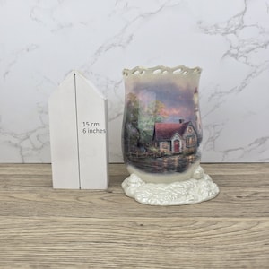 Può includere: Un vaso decorativo in ceramica con una scena di faro e cottage, alto circa 15 cm. Il vaso ha un bordo smerlato e una base strutturata. L'immagine sul vaso raffigura un paesaggio sereno con un faro e un cottage.