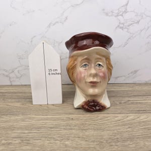 May include: A ceramic mug shaped like a man's head, with a brown hat and a white collar. The mug is 6 inches tall. The man has rosy cheeks and light brown hair. The mug is on a wooden surface.