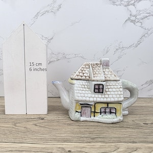 Può includere: Una teiera in ceramica a forma di casa, con facciata bianca e gialla, tetto marrone e camino. La teiera è alta circa 15 cm. La teiera a forma di casa ha un manico e un beccuccio.