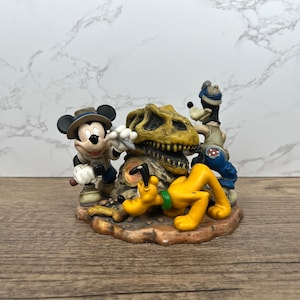 以下が含まれることがあります： ミッキーマウス、グーフィー、プルートが恐竜の頭蓋骨を調べているディズニーの置物。ミッキーはサファリの衣装、グーフィーはブルージーンズ、プルートは横たわっています。置物は茶色の台座に置かれています。
