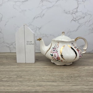 Könnte beinhalten: Eine weiße Keramik-Teekanne mit floralen und goldenen Akzenten. Die Teekanne hat einen Deckel und einen gebogenen Ausguss. Ein weißes, hausförmiges Objekt befindet sich neben der Teekanne. Das hausförmige Objekt ist 15 cm hoch.